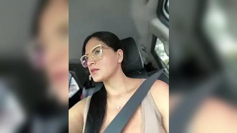 Media: Video of a woman with long dark hair, wearing glasses, beige top, and seatbelt, driving a car. Blurred background, suggesting focus on her.