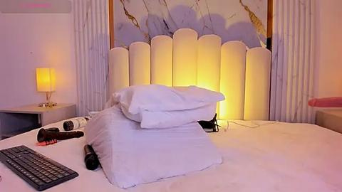 Media: Video of a modern, minimalist bedroom with a plush white bed, white pillows, a gold-leaf headboard, and cream curtains. A laptop, remote control, and a glass of water are on the bed.