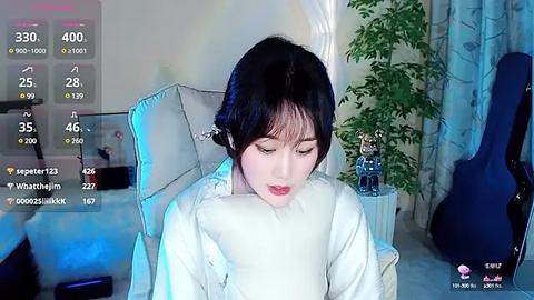 Media: Video of a young East Asian woman with fair skin and black hair, wearing a white sweater, sitting on a white chair in a cozy room. The room includes a guitar, plants, and blue curtains.