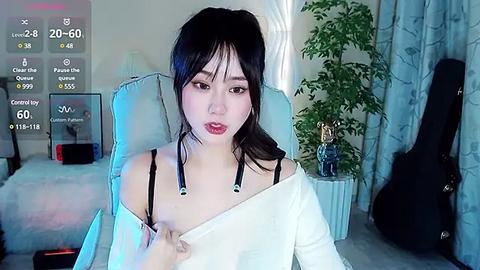 Media: Video of a young East Asian woman with long black hair and pale skin, wearing a white off-shoulder top, sitting in a room with a guitar and potted plant.