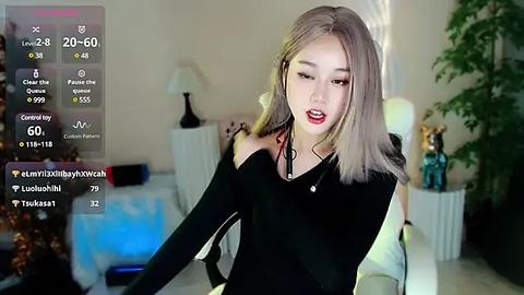 Media: A video of a young Asian woman with long, straight, platinum blonde hair, wearing a black off-shoulder top, seated on a white chair, in a modern, softly lit living room.