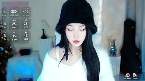 Media: Video of a young woman with pale skin, long black hair, and a black bucket hat, wearing a white, off-shoulder sweater, standing in a softly lit, modern bedroom with a Christmas tree and decorative bottles.