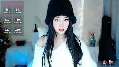 Media: Video of a young Asian woman with long black hair, wearing a black beanie and white off-shoulder sweater, standing in a modern, minimalist room with a Christmas tree and a guitar in the background.