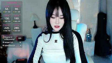 Media: A video of a young East Asian woman with long black hair, wearing a white Chanel jacket, sitting in a dimly lit bedroom with a guitar and lamp.