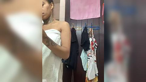 Media: Video of a woman in a bathroom with a towel covering her chest, holding a phone, with clothes hanging on a rack in the background.