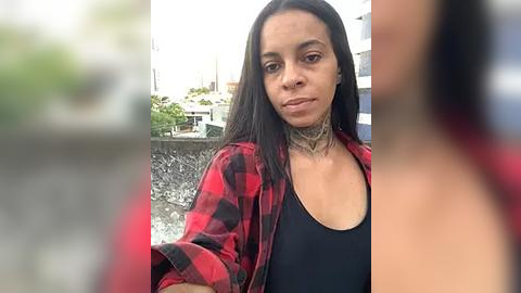 Media: Video of a woman with medium brown skin, straight black hair, wearing a black tank top and a red and black plaid shirt. She has a tattoo on her neck and is standing outdoors next to a concrete wall with greenery.