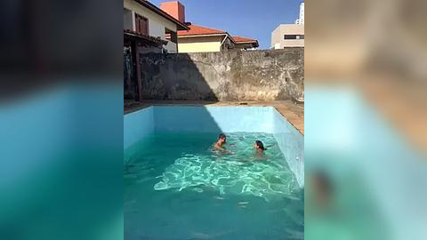 Media: Video of two young boys in a small, blue-tiled, rectangular pool. One boy is in the water, while the other is on the edge. Background features a concrete wall, a red-tiled roof, and a clear blue sky.