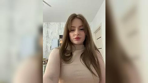 Media: Video of a young Caucasian woman with long, light brown hair, fair skin, and a slim figure, wearing a beige turtleneck sweater. Background features a modern, bright room with white walls, marble accents, and a partially visible mirror.