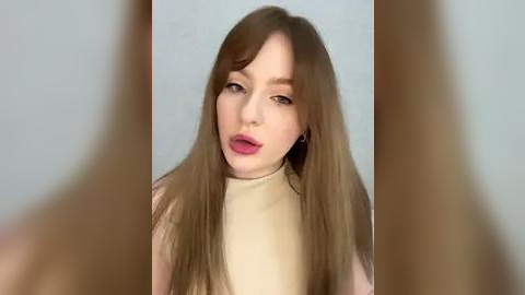 Media: A video of a young, fair-skinned woman with long, straight, light brown hair, wearing a beige turtleneck sweater. She has a neutral expression, with light makeup and pink lipstick. The background is plain and out of focus.