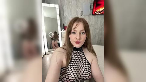 Media: Video of a fair-skinned, red-haired woman in a black, fishnet halter top, sitting in a modern bathroom with a lit fireplace and a large mirror.