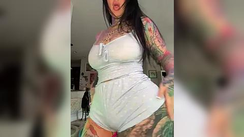 Media: Video of a tattooed woman in a light blue crop top and matching shorts, posing provocatively with her tongue out. Background includes a blurred, modern living room.