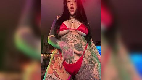 Media: Video of a tattooed woman in a red bikini, posing provocatively with tongue out, in a dimly lit room with colorful, blurred figures in the background.