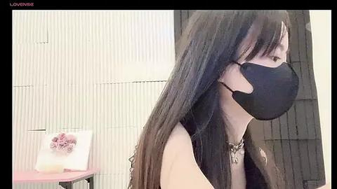 Media: Video of a woman with long black hair, wearing a black mask and necklace, seated in a room with white vertical blinds, pink vase, and a framed picture.
