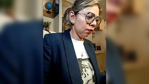 Media: Video of a woman with blonde hair in glasses, wearing a dark blazer over a white shirt with a graphic print. Background features a globe, plants, and abstract art.