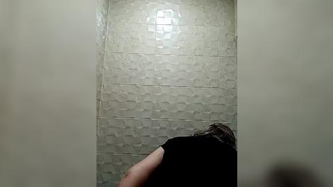 Media: Video of a person with shoulder-length brown hair, wearing a black shirt, standing in a dimly lit, beige-tiled bathroom with a textured pattern.