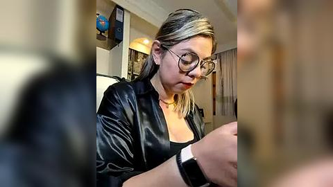 Media: A video of a blonde woman with glasses and a black leather jacket, seated indoors, wearing a smartwatch, looking intently at a phone.