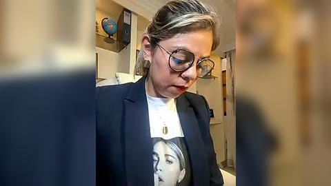 Media: Video of a woman with blonde hair in glasses, wearing a navy blazer over a white t-shirt with a Marilyn Monroe print, sitting in a cluttered room with a globe and books on shelves.