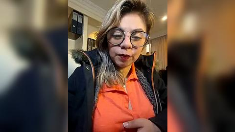 Media: Video of a middle-aged woman with glasses, wearing a bright orange blouse and a black jacket with fur trim, speaking earnestly in a dimly lit room.