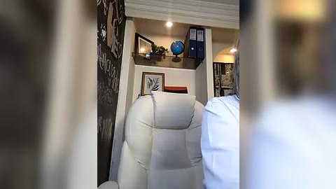 Media: Video of a cozy home office with a white leather recliner chair, a small bookshelf, and a globe on a shelf, surrounded by warm lighting, framed artwork, and a wall covered with decorative elements.