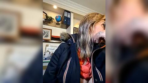 Media: Video of a woman with long, light brown hair wearing a dark blue jacket and red shirt, standing in a modern office with shelves displaying framed certificates and a globe.