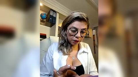 Media: Video of a Latina woman with light skin, short wavy hair, wearing glasses and a white lab coat, examining a small object in a modern office setting.