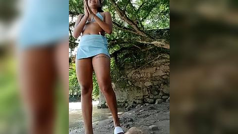 Media: Video of a slender woman with tanned skin and long dark hair, wearing a light blue bikini top and high-waisted shorts, standing on a rocky beach surrounded by lush green trees and a rustic stone wall.