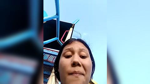 Media: Video of a smiling young Asian woman with dark hair, wearing a blue and white striped shirt, perched on a blue metal playground structure, looking up at the sky.