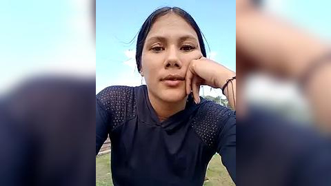 Media: Video of a young woman with light brown skin and straight black hair, wearing a black long-sleeve shirt, leaning on her hand in a grassy outdoor setting.