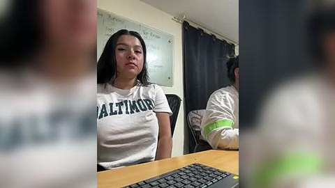 Media: Video of a young woman with long dark hair, wearing a white \"Valencia\" t-shirt, sitting at a wooden desk with a black keyboard. Background includes a whiteboard with notes, a dark curtain, and a man in a striped shirt.