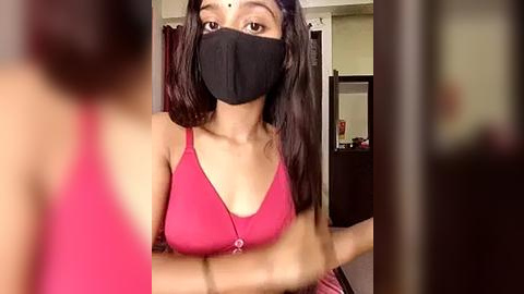 Media: Video of a young South Asian woman with medium skin tone, wearing a pink spaghetti-strap top, black face mask, and a forehead bindi. She's indoors, in a dimly lit room with a blurred background.