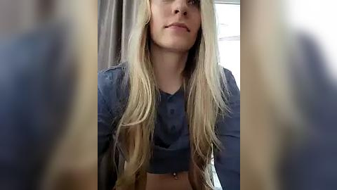 Media: A video of a young, fair-skinned woman with long, straight blonde hair, wearing a blue crop top, smiling slightly, in a bright room with windows and light-colored curtains.