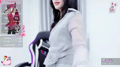 Media: A video of a fair-skinned woman with long black hair and red lipstick, wearing a grey blazer and black stockings, standing in a modern room with a gaming chair.