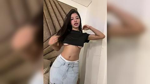 Media: Video of a young woman with long black hair, wearing a black crop top, light blue high-waisted jeans, and white underwear, posing confidently indoors with beige curtains in the background.