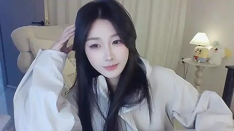 Media: Video of an East Asian woman with long black hair, fair skin, and a slender physique, smiling in a cozy room with a white armchair, a nightstand with a lamp, and a plush toy.