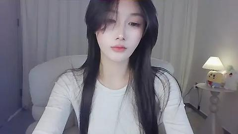 Media: A video of an East Asian woman with long black hair, fair skin, and a slender build, wearing a white long-sleeved shirt, seated in a dimly lit room with white curtains and a lamp.