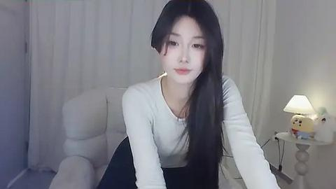 Media: Video of a young Asian woman with long black hair, fair skin, and large eyes, wearing a white long-sleeve top and a black skirt, leaning forward, in a softly lit, minimalist room with white walls and a lamp.