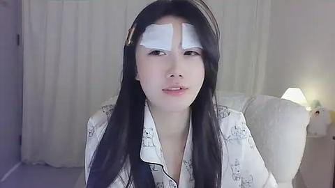 Media: Video of a young woman with long black hair, wearing a white shirt with cartoon characters, and white bandages over her eyes, sitting in a room with white curtains and a bedside lamp.