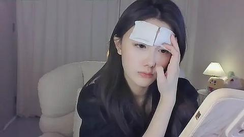 Media: A video of an Asian woman with long black hair, wearing a black shirt, and a white bandage on her forehead, sitting in a dimly lit room with a white armchair, a lamp, and stuffed toys.