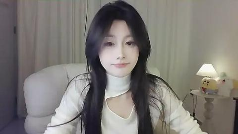 Media: Video of an East Asian woman with long black hair, wearing a white turtleneck, sitting in a modern, softly-lit room with a beige armchair and a lamp on a white nightstand.