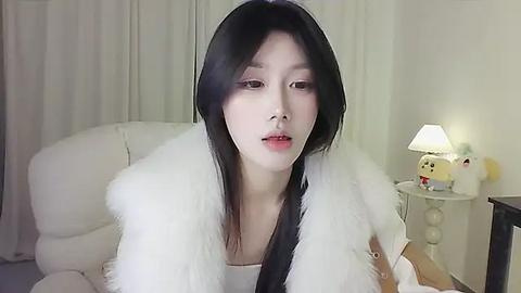 Media: Video of a young East Asian woman with long, straight black hair, wearing a white fur coat, looking serious, in a minimalist bedroom with a white bed, nightstand, and soft lighting.