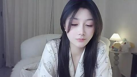 Media: Video of a young East Asian woman with long, straight black hair, wearing a white robe with light gray patterns. She has a calm expression. Background features a white couch, a nightstand lamp, and stuffed toys.