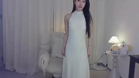 Media: Video of an East Asian woman in a white, sleeveless, halter-neck dress, standing in a softly lit, minimalist living room with white curtains, a white armchair, and a table lamp.