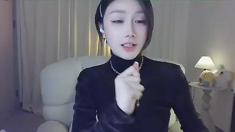 Media: Video of an Asian woman with pale skin, dark hair, wearing a black leather jacket, standing in a dimly lit room with a white chair, a lamp, and a stuffed animal.