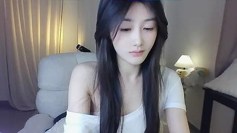 Media: A video of an East Asian woman with long black hair, wearing a white off-shoulder top, sitting on a beige couch. Soft lighting from a table lamp in the background.