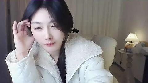 Media: A video of a young Asian woman with shoulder-length black hair, wearing a white, fluffy jacket, adjusting her hair in a cozy, softly lit room with a lamp and a teddy bear.