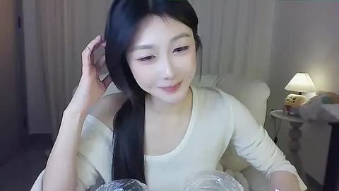 Media: Video of an East Asian woman with long black hair, light skin, and a slim figure, wearing a cream sweater. She sits on a white couch in a dimly lit room with a table lamp, stuffed toys, and sheer white curtains in the background.