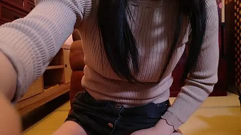 Media: Video of an Asian woman with long black hair, wearing a ribbed beige sweater and black denim shorts, sitting on a yellow mat in a wooden room with bookshelves and a red door in the background.