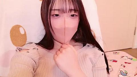 Media: Video of an Asian woman with long black hair, light skin, and a white mask, sitting on a bed with a patterned sheet, holding a finger to her lips.