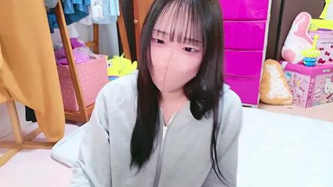 Media: Video of a young Asian woman with long black hair, wearing a light blue hoodie, sitting on a white floor in a cluttered room with colorful toys, clothes, and a pink bag in the background.