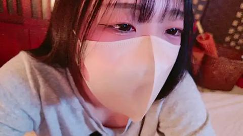 Media: Video of an Asian woman with straight black hair, wearing a light blue shirt and a white mask, looking down. Background includes a red cushion and a brown teddy bear.